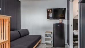 Huoneessa on musta sohva, seinälle asennettu televisio, musta jääkaappi ja puinen kaappi.
