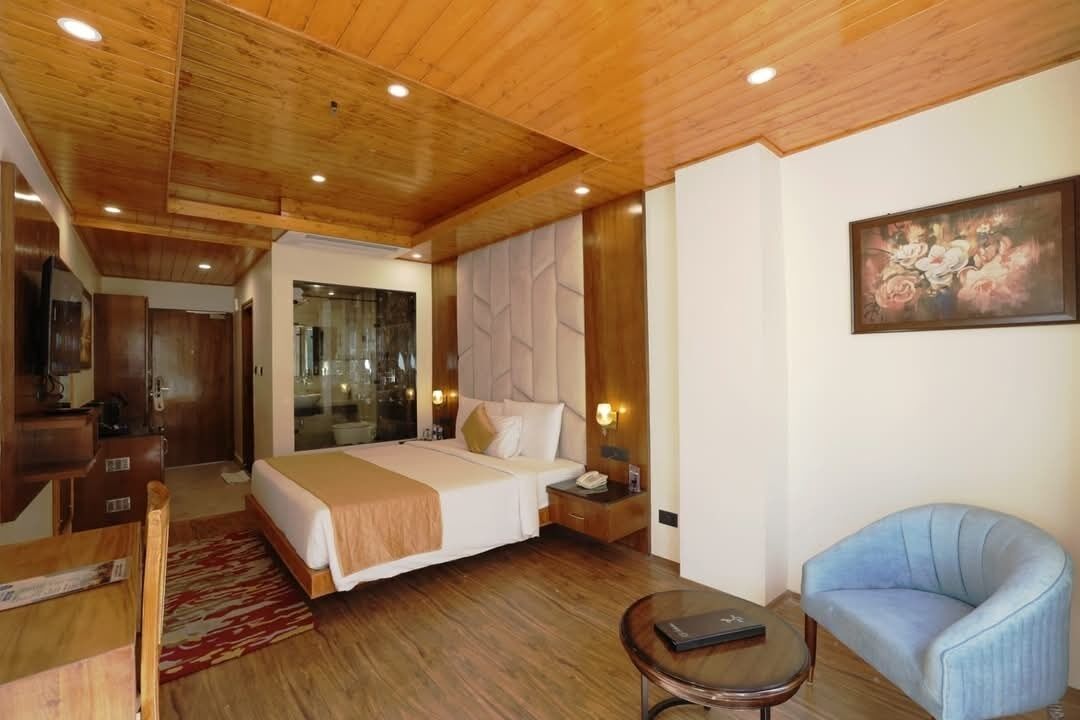 A hotel room with a wooden ceiling, a bed with a beige bedspread, a nightstand with a lamp, a blue armchair, and a painting on the wall.