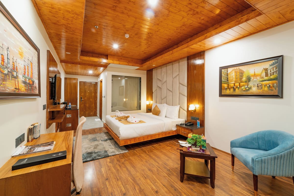 A hotel room with a wooden ceiling, a large bed, a desk with a chair, a small table with a plant, and a painting on the wall.