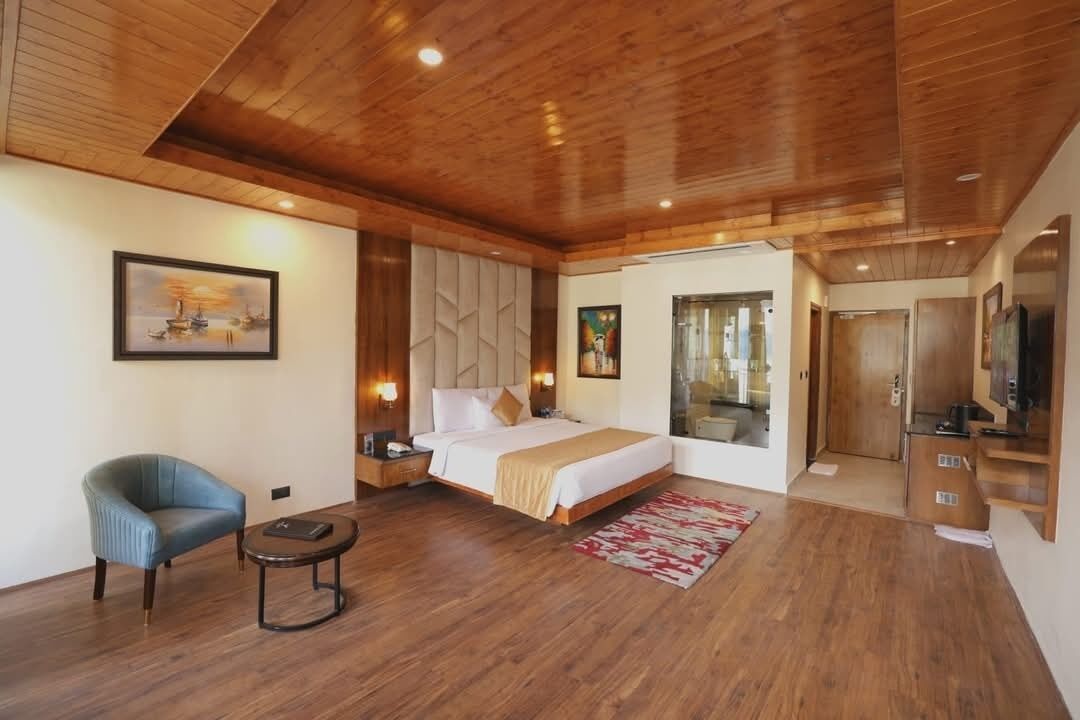 A spacious bedroom with a wooden ceiling, a large bed, a blue armchair, a small round table, and a large window.