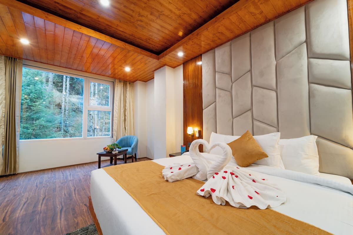 A hotel room with a large bed, a wooden ceiling, a window with a view of trees, a blue chair, and a nightstand with a lamp.