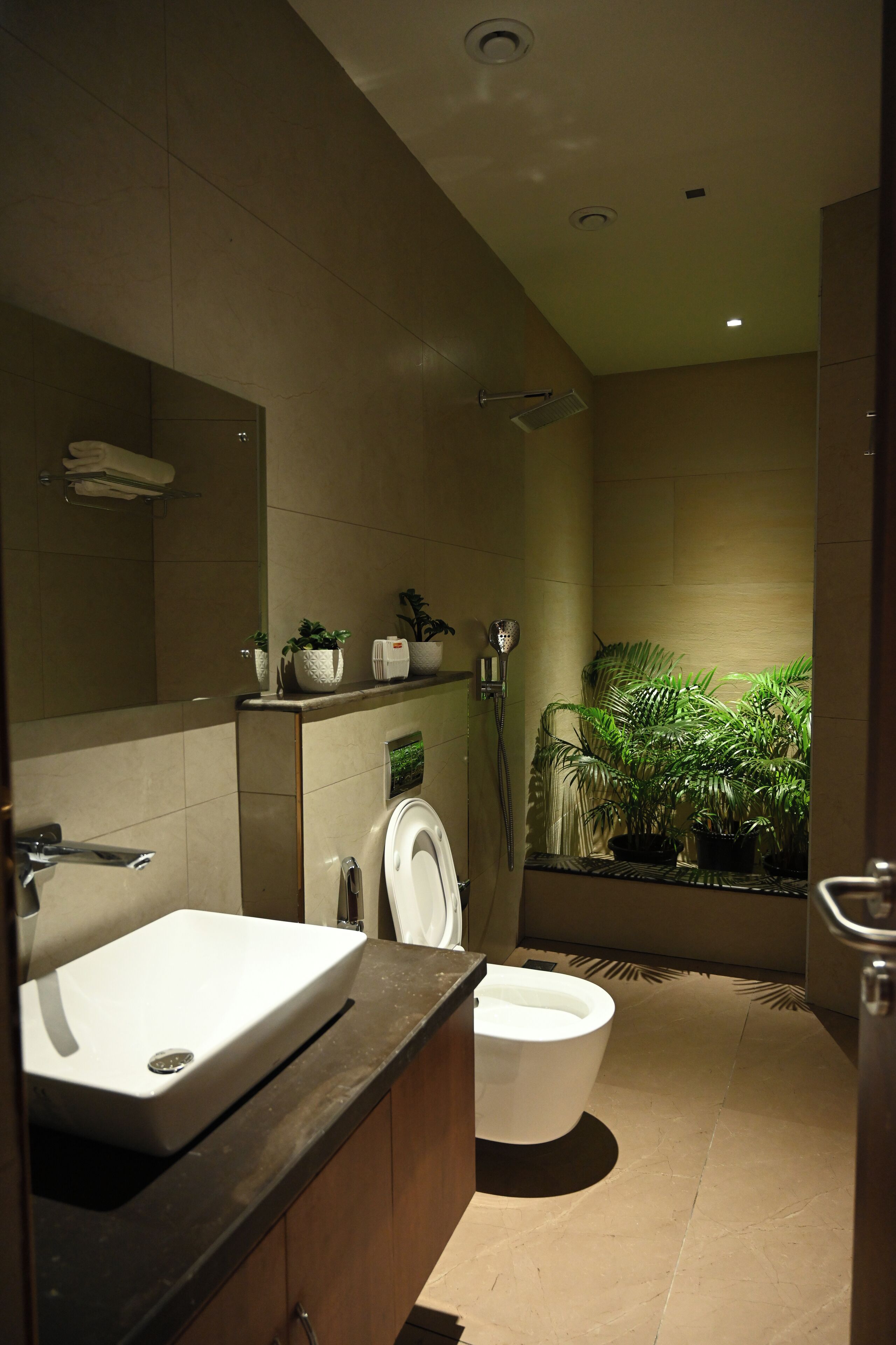 A modern bathroom with a sink, toilet, and shower. There are plants on the shelves and a mirror above the sink.
