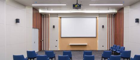 A conference room with rows of blue chairs, a large projection screen, and a whiteboard.