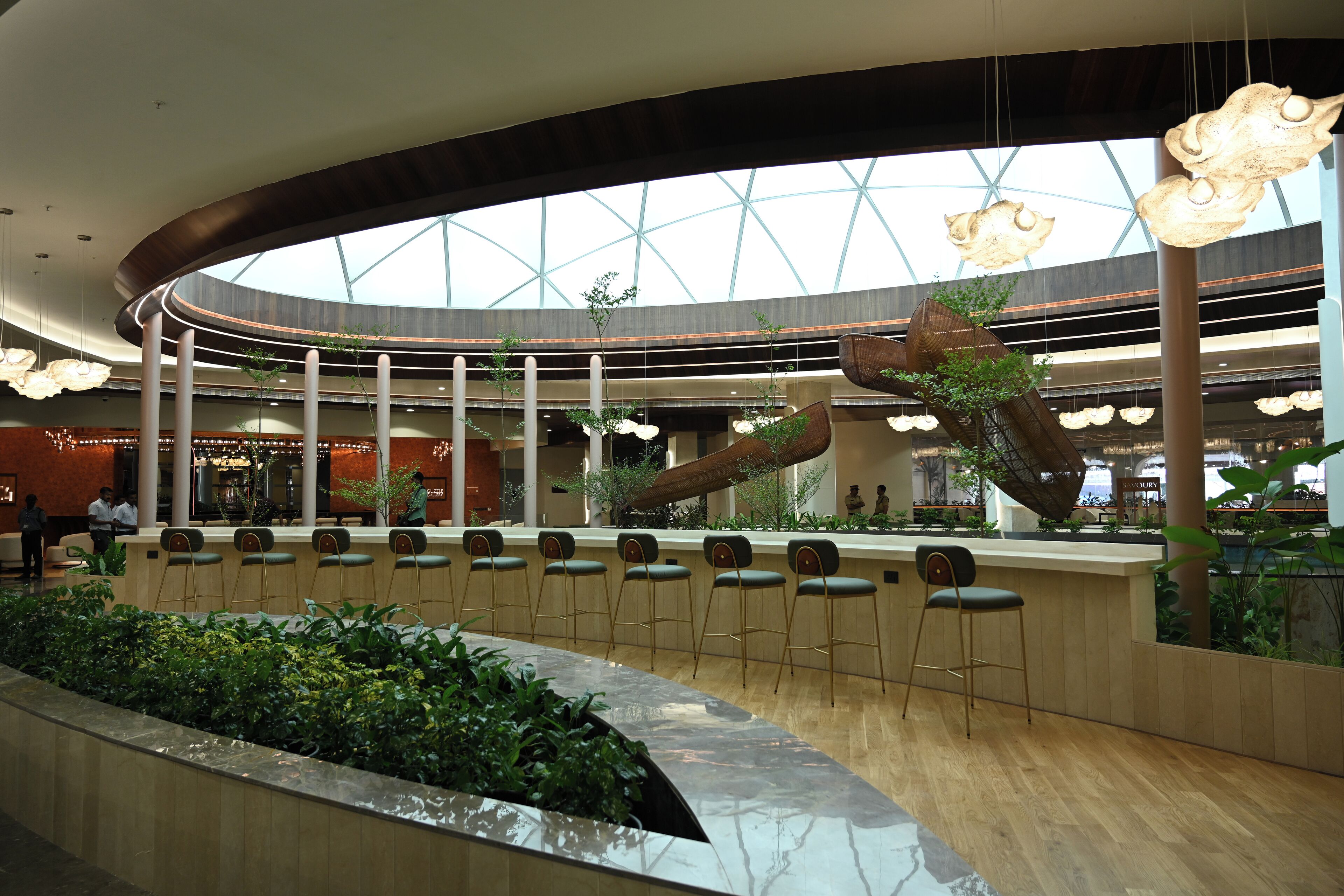 A modern indoor space with a curved ceiling, a central bar area with stools, and a variety of plants.