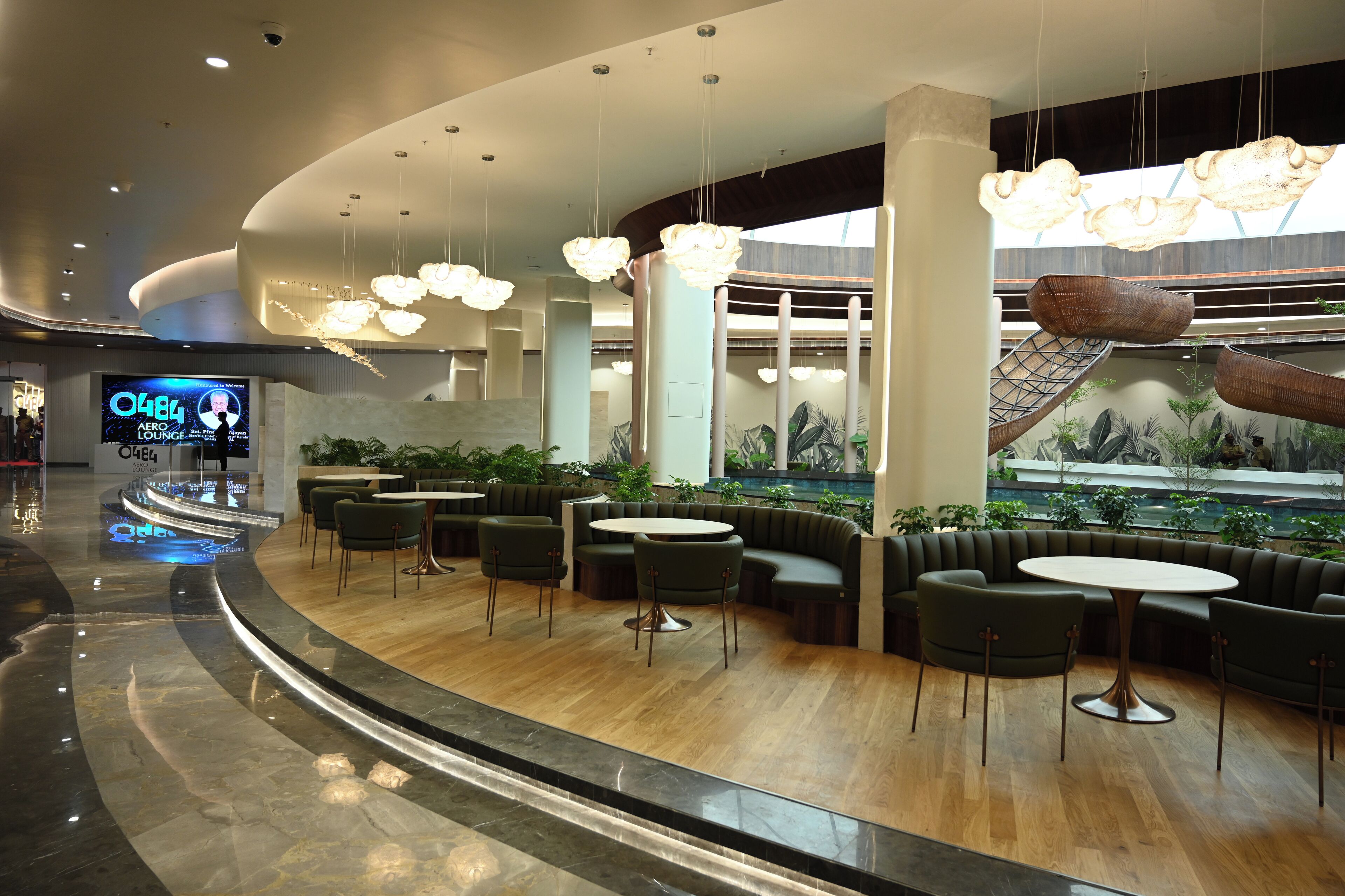 A modern lounge area with round tables and green sofas, a large digital screen, and a central water feature.