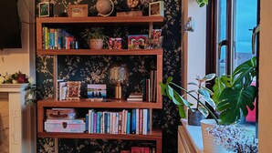 Ein Zimmer mit Bücherregal, voll mit Büchern und Dekoration, einem Klavier und einem Fenster mit Pflanzen.