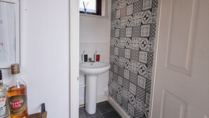 Ein Badezimmer mit einem weißen Waschbecken, einer Wand mit Muster und einem Fenster. Auf einem Regal stehen Flaschen und eine Tür mit Fußmatte.