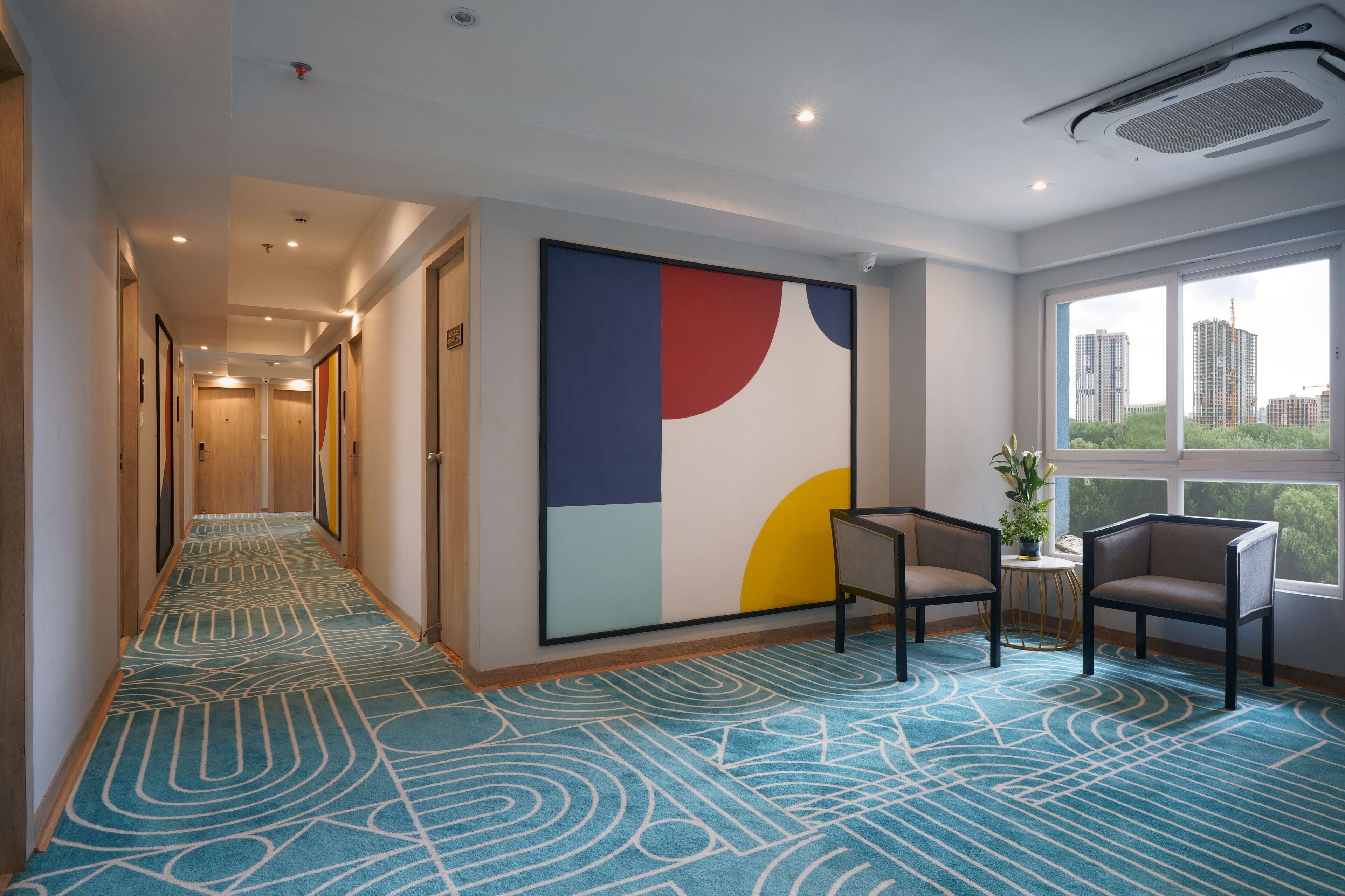 Un pasillo de hotel con alfombra estampada, un gran cuadro abstracto en la pared y una zona de estar con vistas a edificios a través de amplias ventanas.