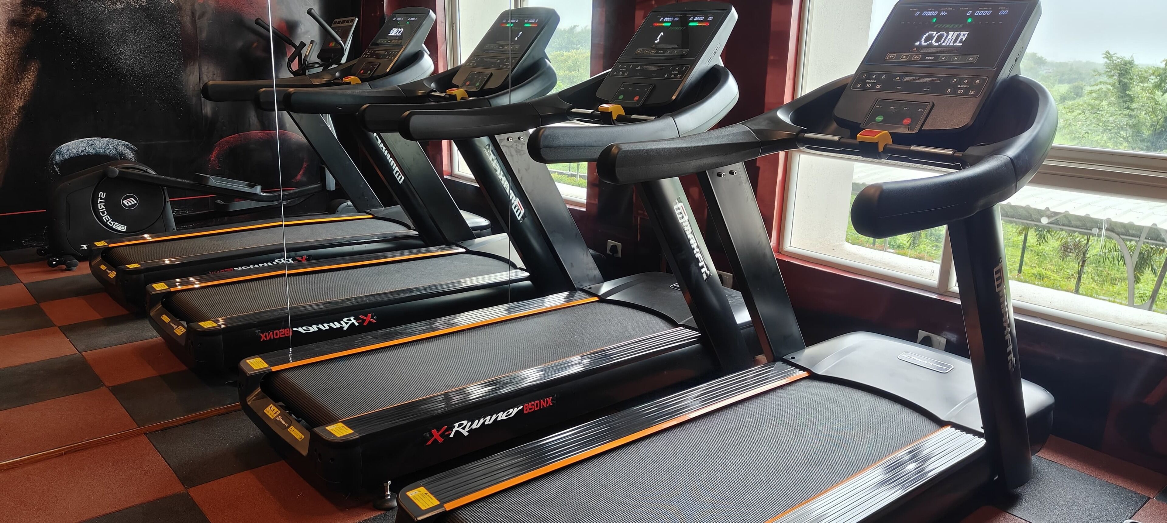 Una hilera de cintas de correr con pantallas digitales, ubicadas en un gimnasio con un patrón de piso a cuadros.