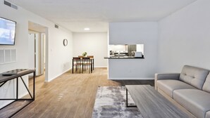 A modern living room with a sofa, a coffee table, and a dining area with chairs and a small plant. - Northcliffe 404 I Fully Furnished 1 Bedroom 1 Ba (Winston-Salem)