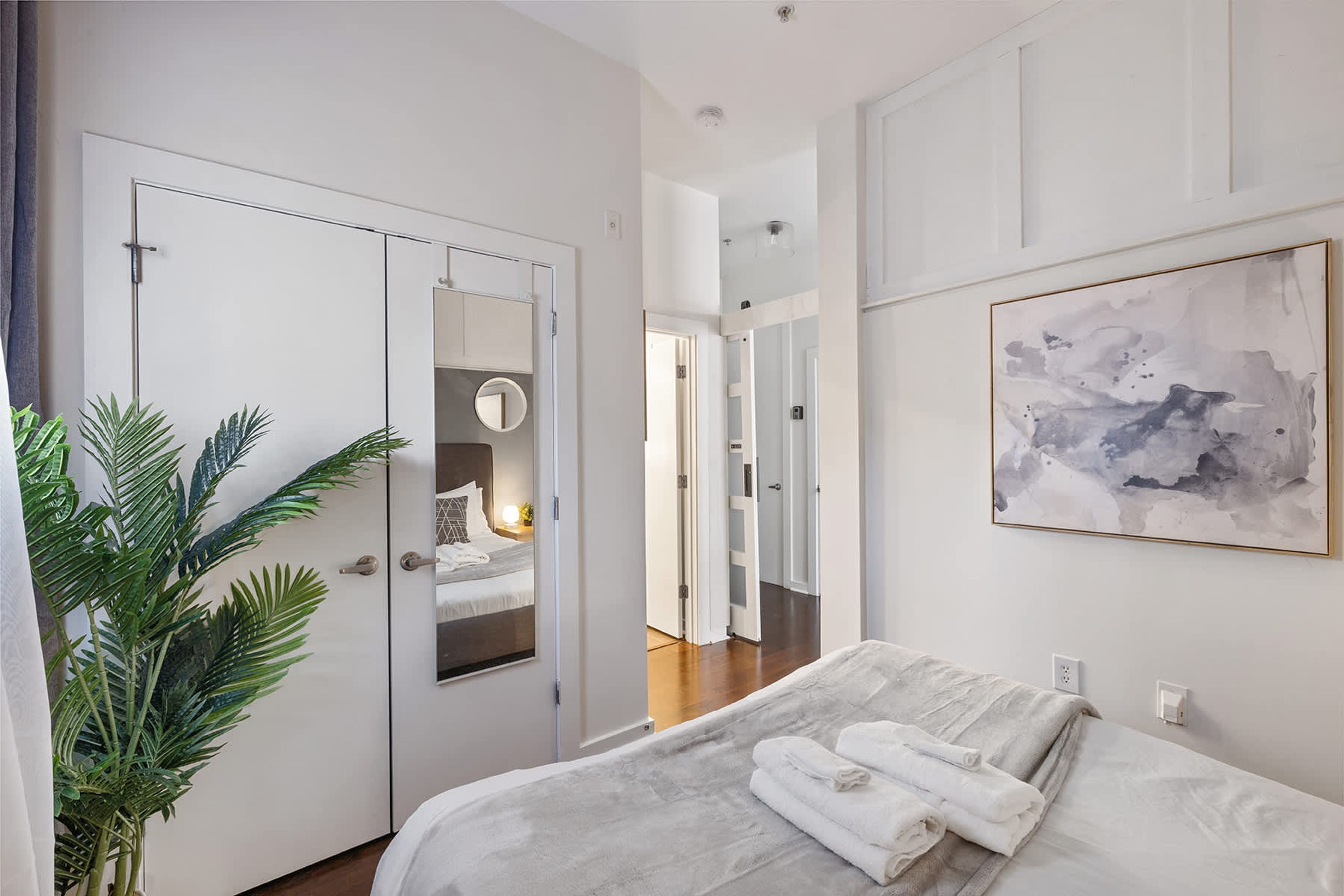 A modern hotel room with a bed, a large mirror, a potted plant, and a painting on the wall.