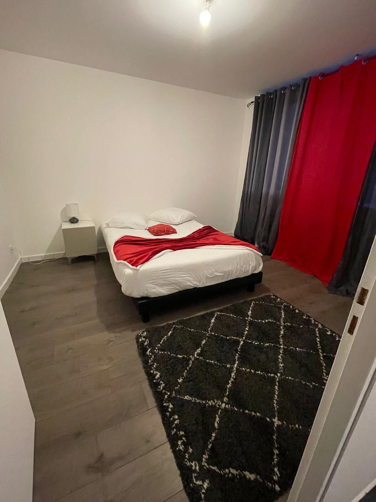 A bedroom with a bed, a nightstand, a red curtain, and a patterned rug.