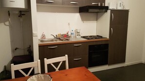 Eine kleine Küche mit Herd, Spüle und Schränken. Ein Tisch mit Stühlen und einem Wasserkocher stehen auf der Arbeitsplatte.
