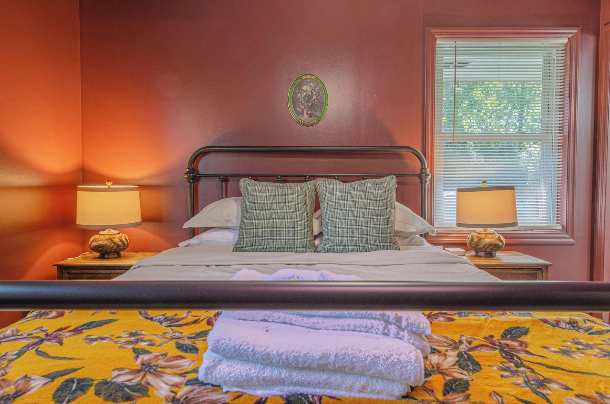 A neatly made bed with two pillows, a window with blinds, and two bedside lamps on wooden nightstands.