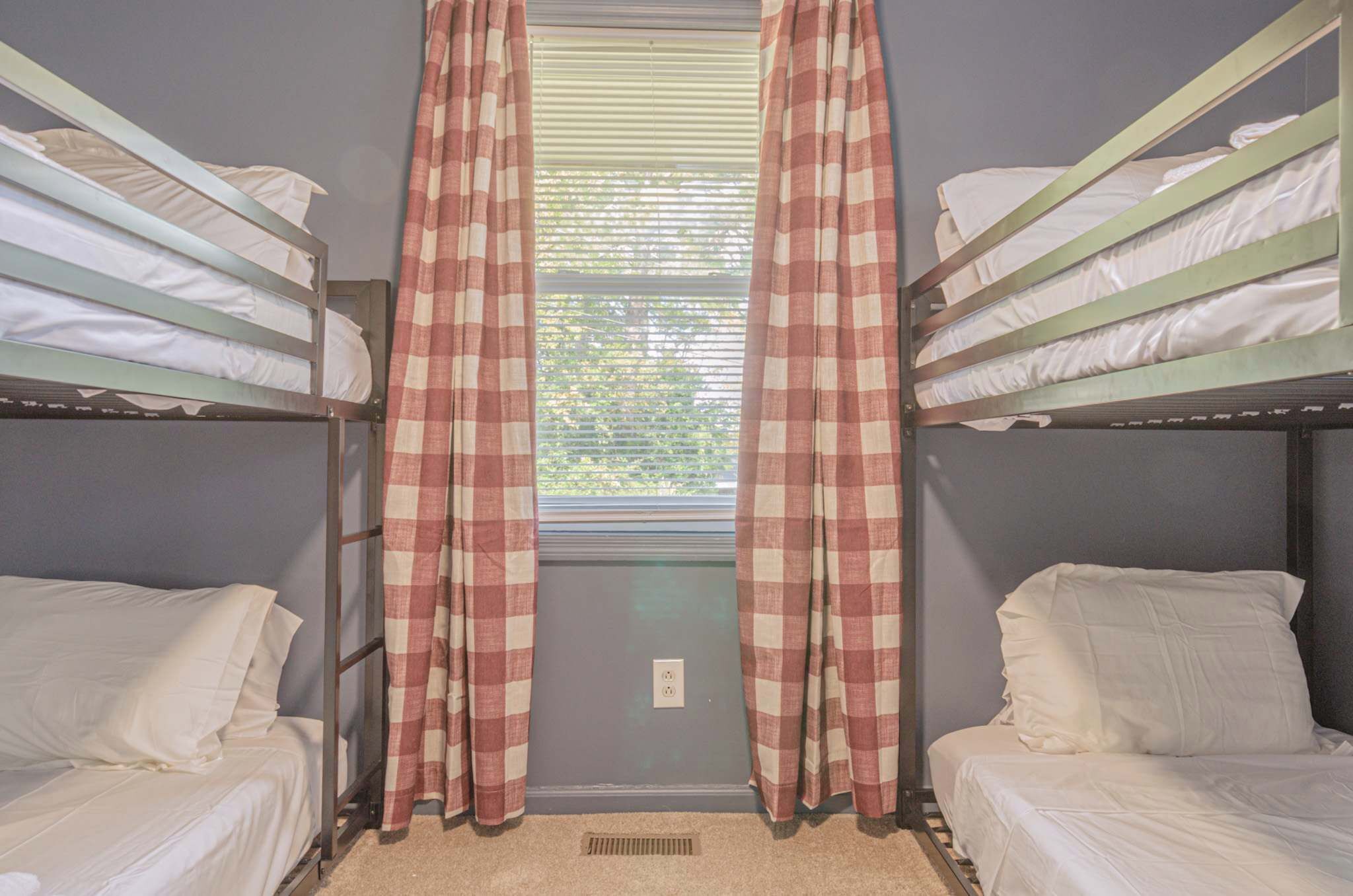 Quarto com duas camas beliche, cortinas xadrez, janela com persianas e uma tomada na parede.