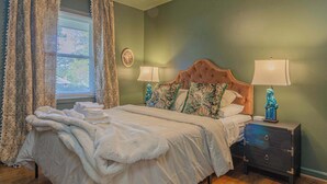 Un lit bien fait, recouvert d’une couverture blanche et moelleuse, avec deux oreillers assortis et une tête de lit. On y voit deux tables de chevet avec des lampes, et une petite statue sur l’une d’elles. Une fenêtre avec des rideaux à motifs floraux et une horloge murale sont également visibles.