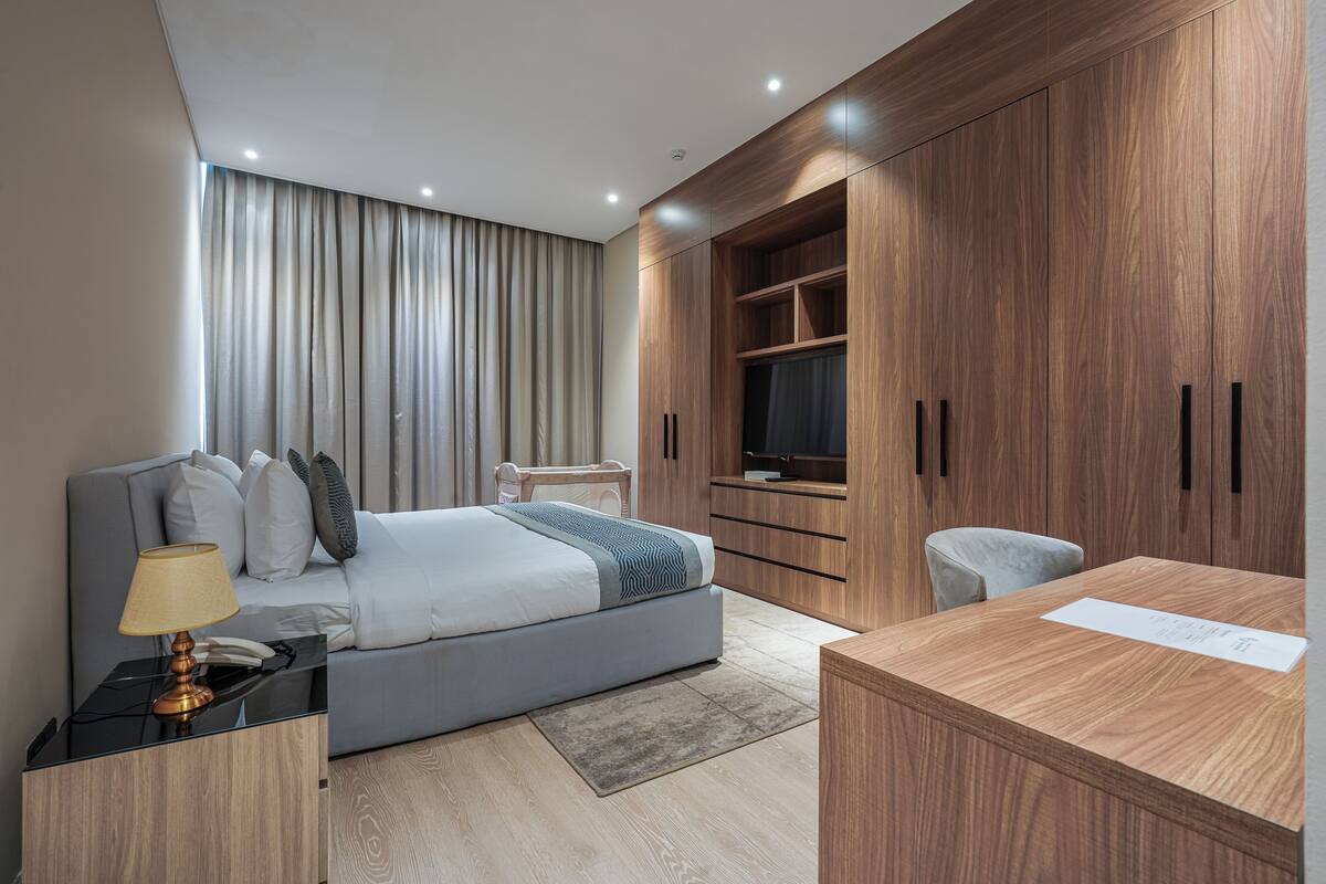 A modern hotel room with a bed, bedside table, lamp, and a wooden desk with a chair.