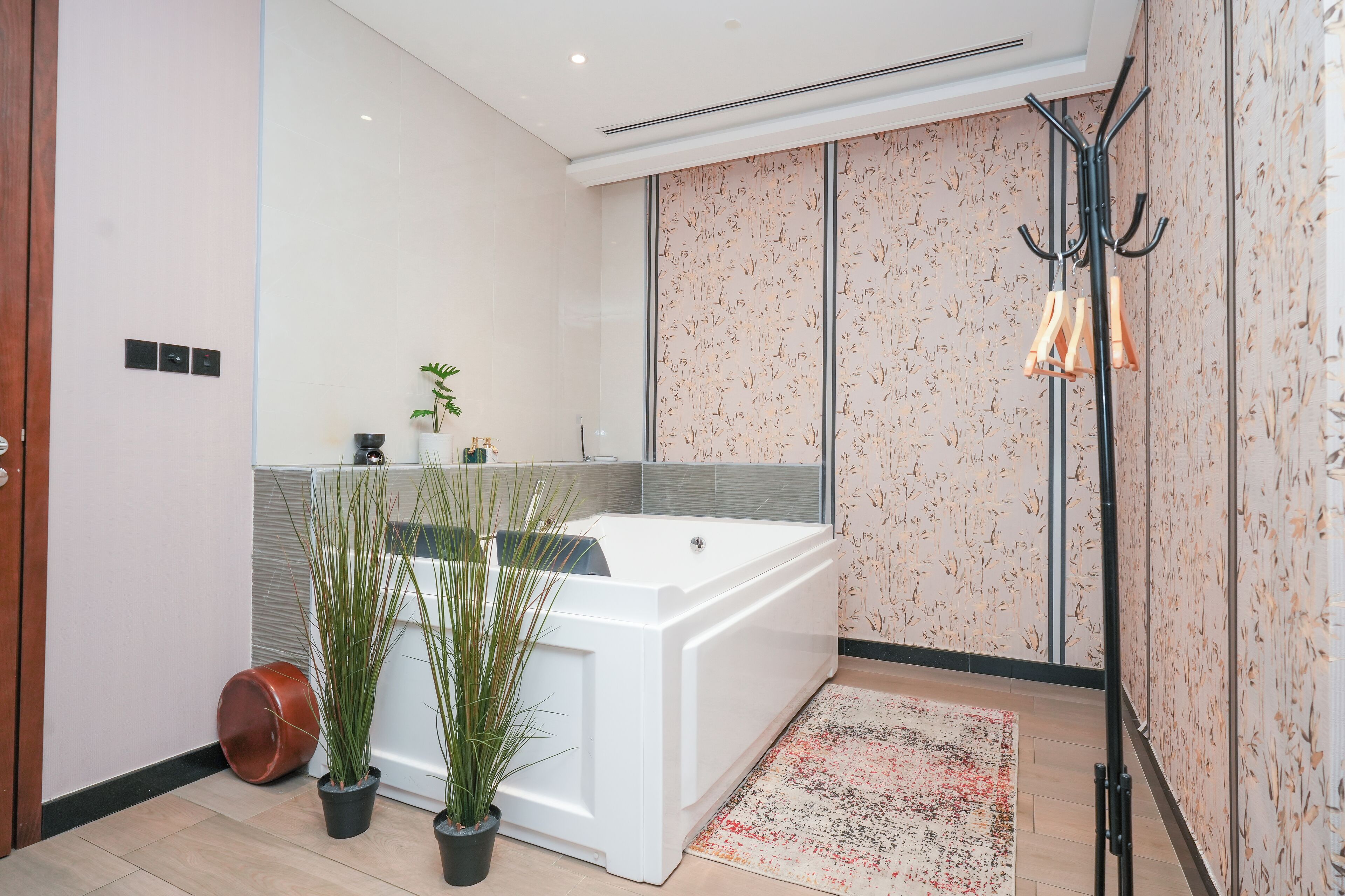 A modern bathroom with a large white bathtub, two potted plants, a wooden floor, and a patterned wall.