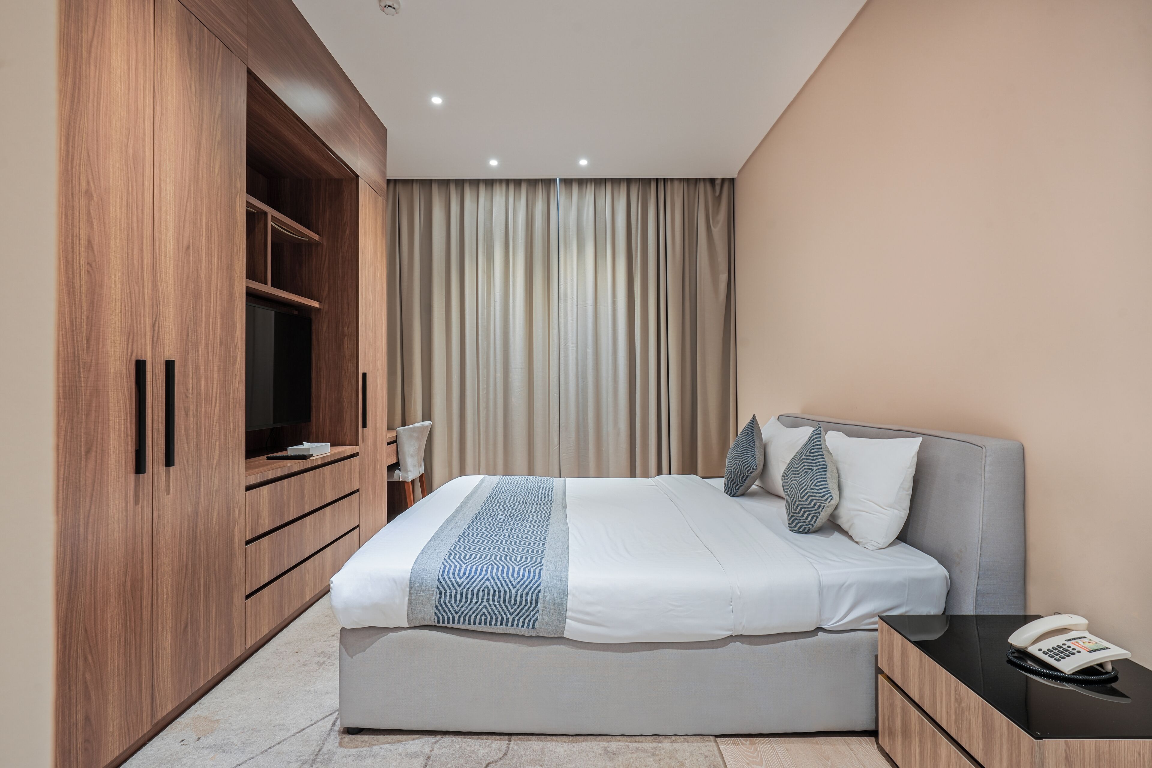 A modern hotel room with a large bed, a wooden wardrobe, a bedside table with a phone, and a curtain backdrop.