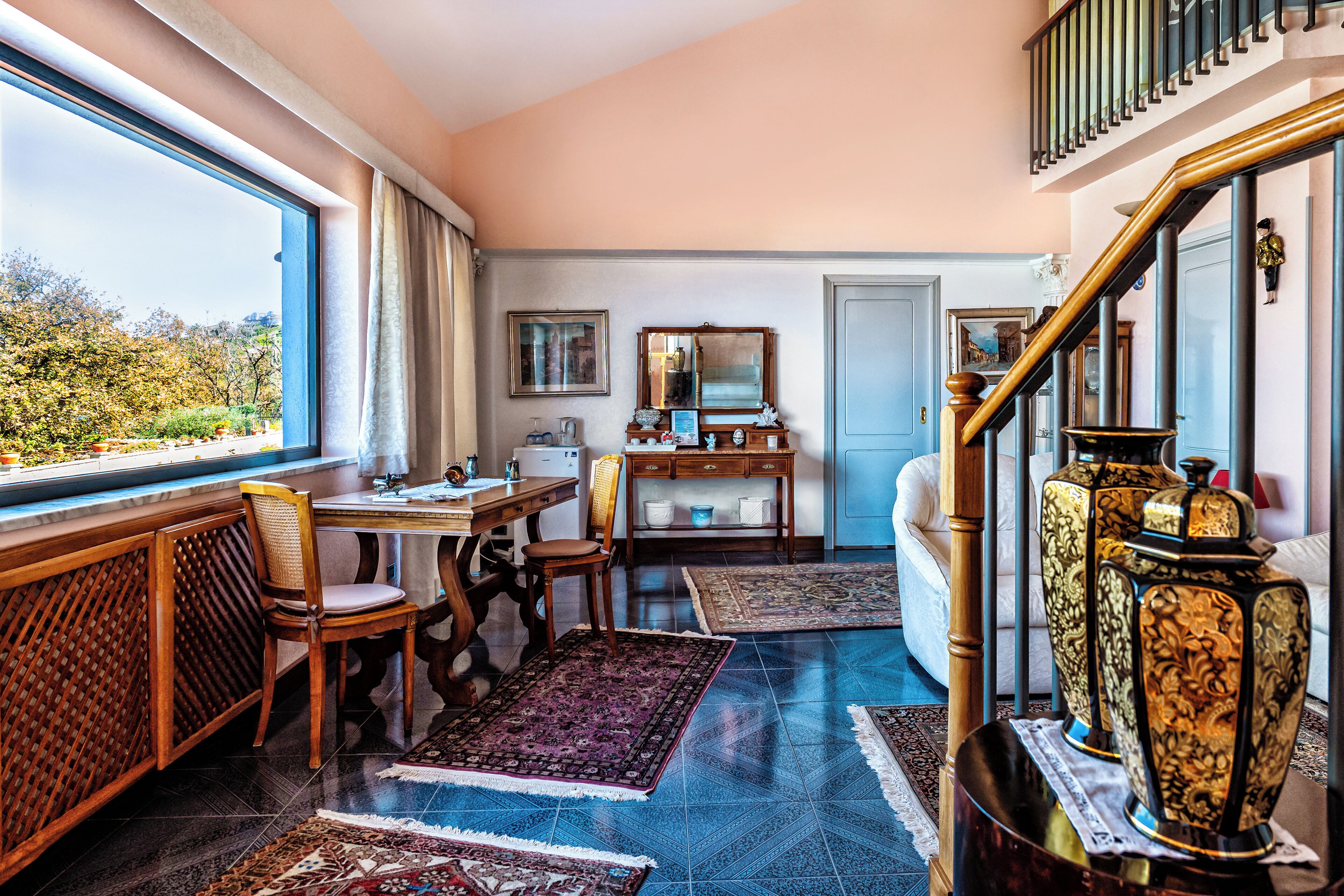 A room with a large window, a wooden desk, a chair, a sofa, and a staircase with a railing.