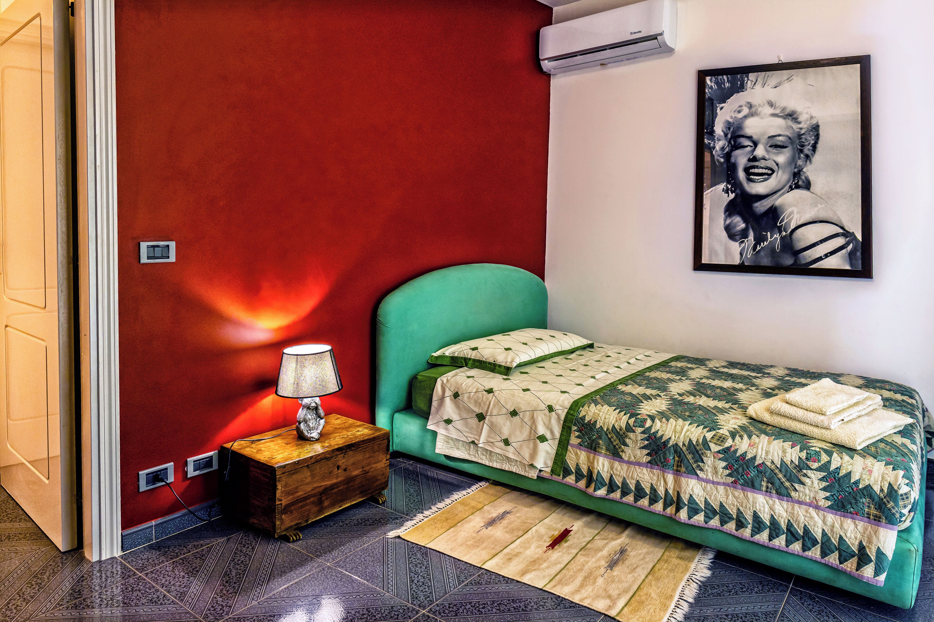 Un dormitorio con cama verde, pared roja, un cuadro enmarcado, mesita de noche con lámpara y alfombra estampada.