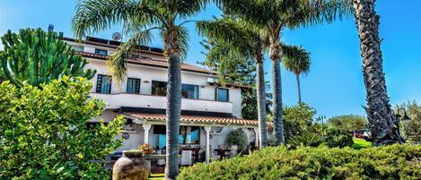 A two-story building with a tiled roof, surrounded by lush greenery including palm trees and various shrubs.
