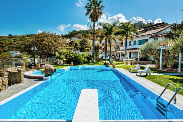 A swimming pool with a clear blue water, surrounded by a stone wall and a grassy area. - Villa Bency (Capo d'Orlando)