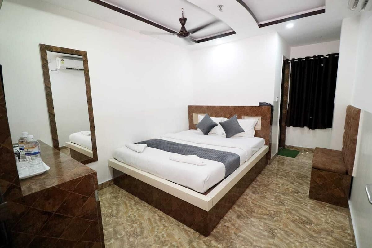 A hotel room with a bed, a bedside table with water bottles, a mirror, a ceiling fan, and a black curtain.