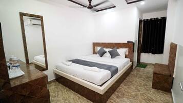 Habitación de hotel con cama, mesita de noche con botellas de agua, espejo, ventilador de techo y cortina negra.