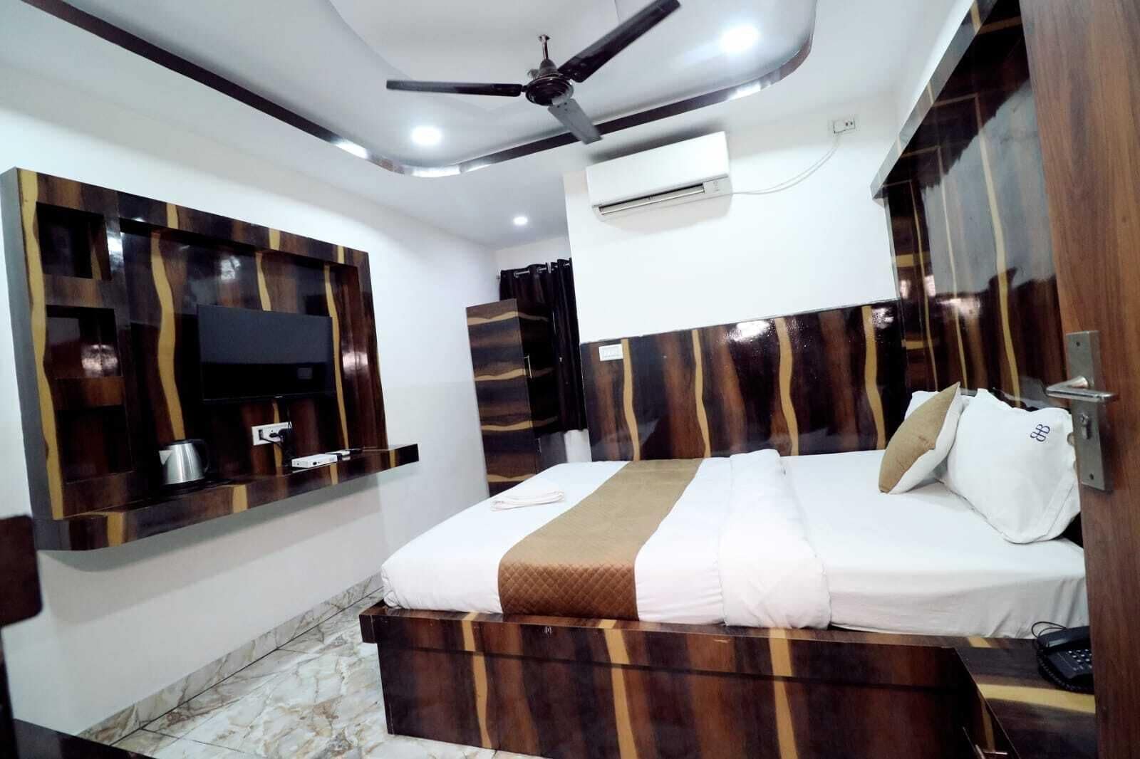 A modern hotel room with a bed, built-in wooden headboard, a flat-screen TV, an air conditioning unit, and a ceiling fan.