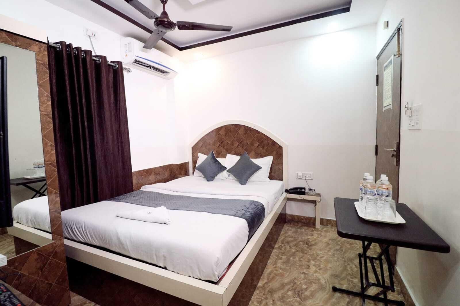 Habitación de hotel con cama, mesita de noche con teléfono y botellas de agua, ventilador de techo y espejo.