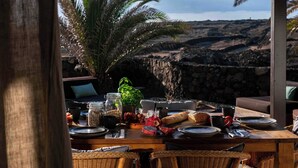 A dining table set with plates, cutlery, and a variety of food items, overlooking a rocky landscape with a palm tree in the background. - Villa by the Sea (Haria)