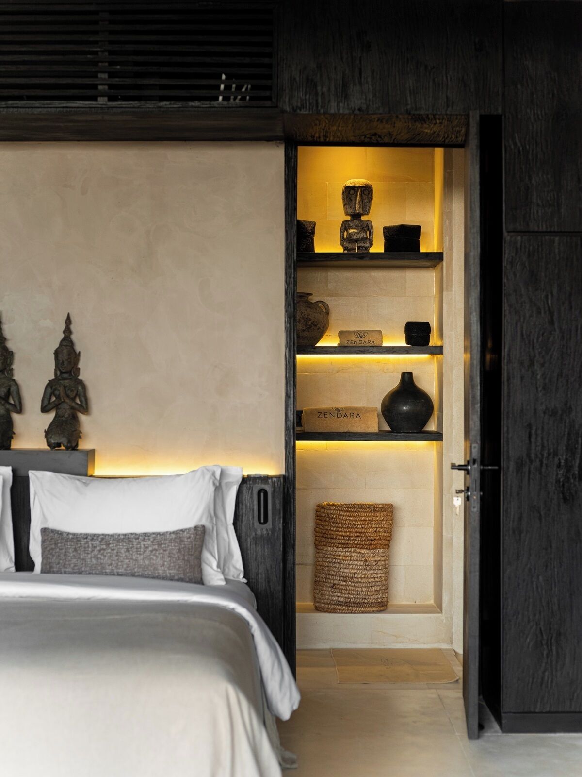 A bedroom with a bed, a nightstand, a shelf with decorative items, and a door leading to another room.