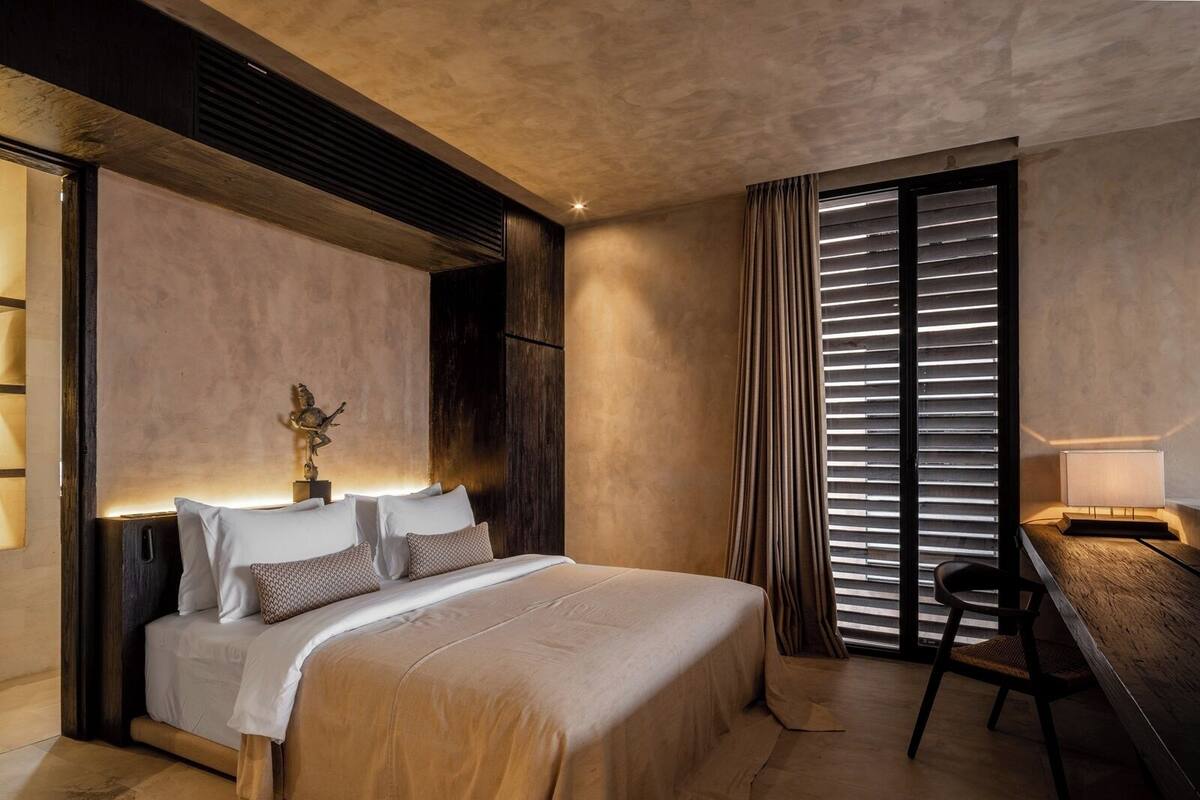 A modern bedroom with a large bed, a desk, and a window with blinds.