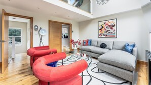 A modern living room with a grey sofa, red armchairs, and a glass coffee table. A large abstract painting and a chandelier are visible. - Vibrant Urban Escape, Family Friendly - Sleeps 8! (London)
