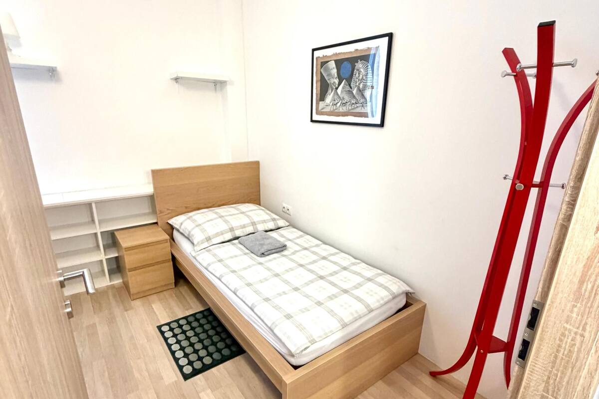 Un dormitorio pequeño y ordenado con una cama individual, una mesita de noche, un pequeño cajón, un cuadro enmarcado en la pared y un perchero rojo.