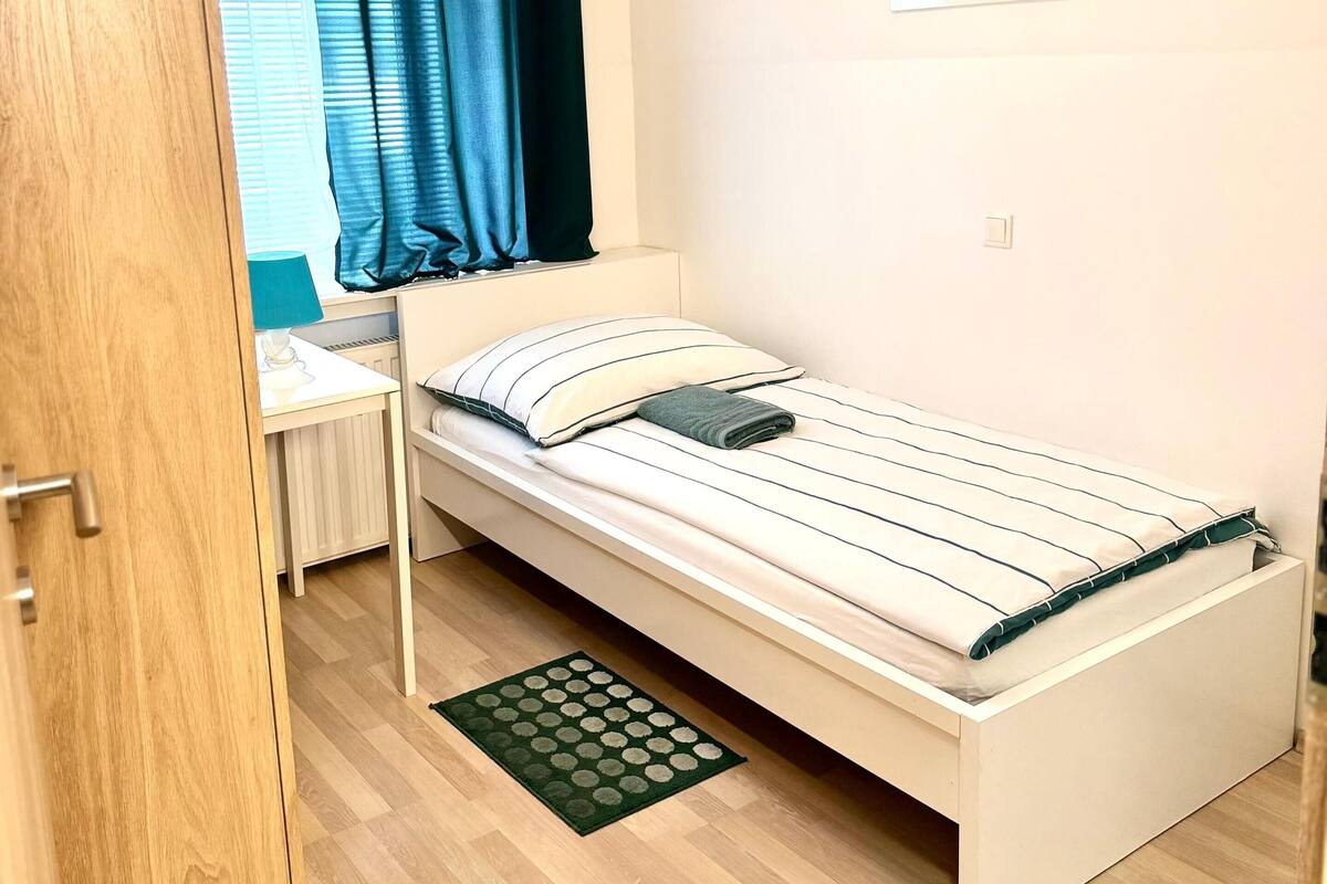 Una cama individual con una colcha a rayas, una mesita de noche, una alfombra pequeña, una ventana con cortinas y una lámpara fijada a la pared.