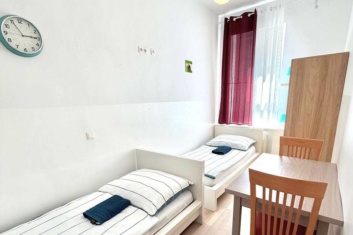 Una habitación pequeña y moderna con dos camas individuales, una silla de madera, una mesa, un reloj y una bombilla.
