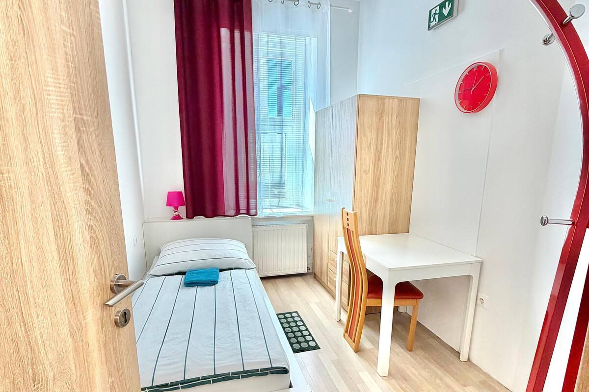 Una habitación con cama, escritorio, silla, ventana con cortinas, un reloj rojo y una lámpara de techo original.