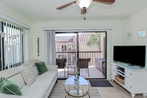 A living room with a sofa, TV, glass coffee table, and a balcony with chairs and a table.