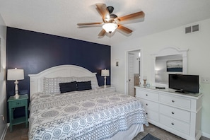 Ein Schlafzimmer mit Bett, Deckenventilator, Kommode, Fernseher und Tür.
