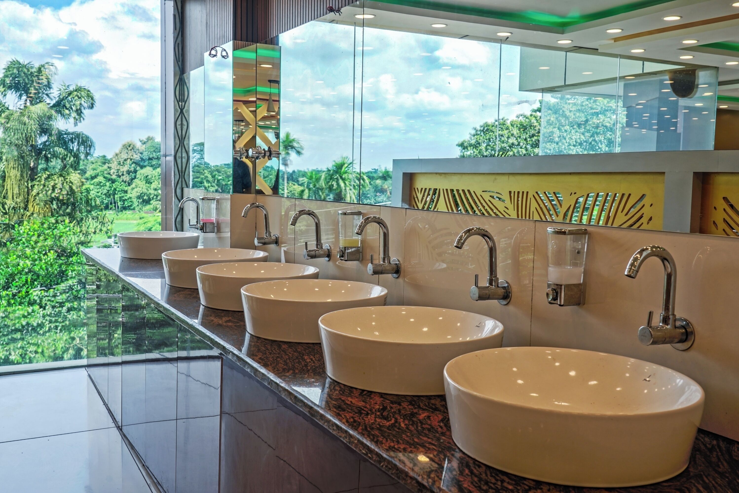 Une salle de bain moderne avec quatre grands lavabos, un grand miroir et une vue sur une végétation luxuriante.