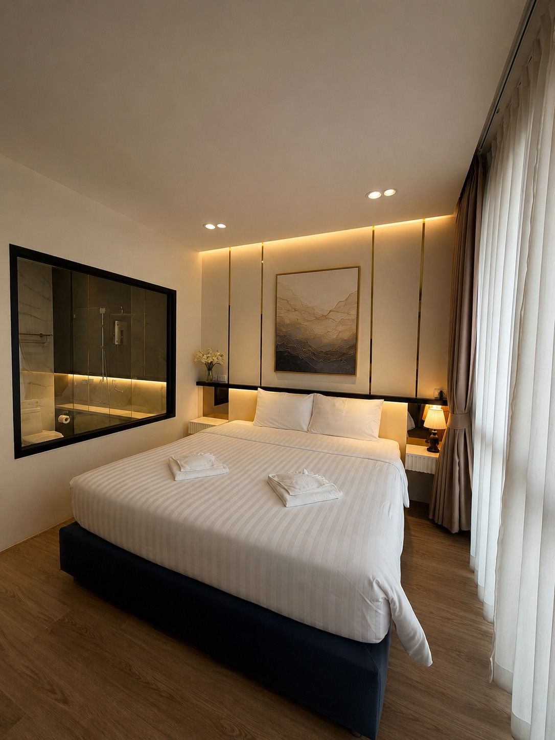 A modern hotel room with a large bed, a mounted artwork, a bathroom visible through a glass partition, and a nightstand with a lamp.
