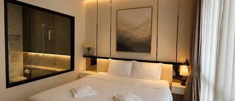 A modern hotel room with a large bed, a mounted artwork, a bathroom visible through a glass partition, and a nightstand with a lamp.