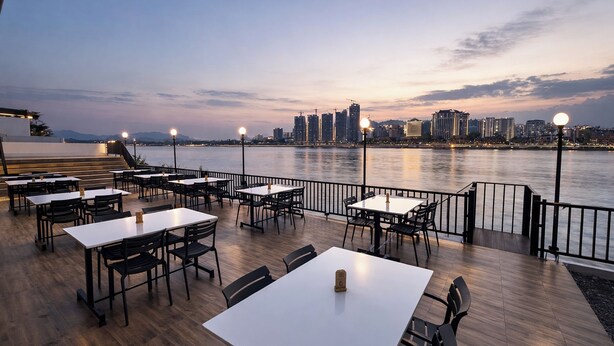 Un área de comedor al aire libre con mesas y sillas, vista al agua y horizonte urbano de fondo.
