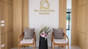 A reception area with a wooden reception desk, two cushioned chairs, a black vase with flowers, and a wall-mounted sign with hotel name. - View khong Hotel (Chiang Saen)