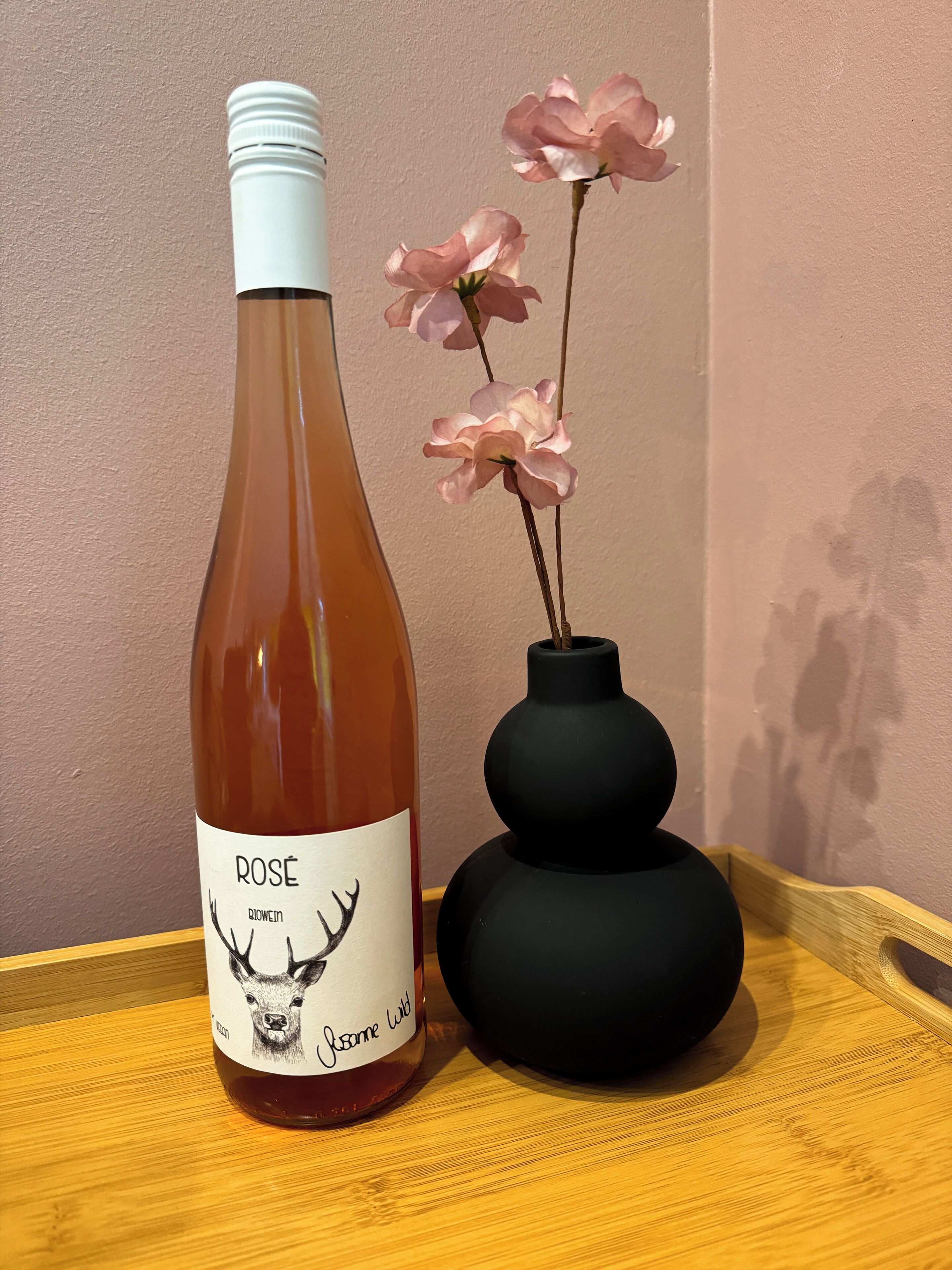 Rosé-viinipullo, jonka etiketissä on peuran kuva ja tekstit »Rose» ja »Bovine Wild». Pullo on asetettu puiseen tarjottimeen mustan maljakon viereen, ja maljakossa on vaaleanpunaisia kukkia.