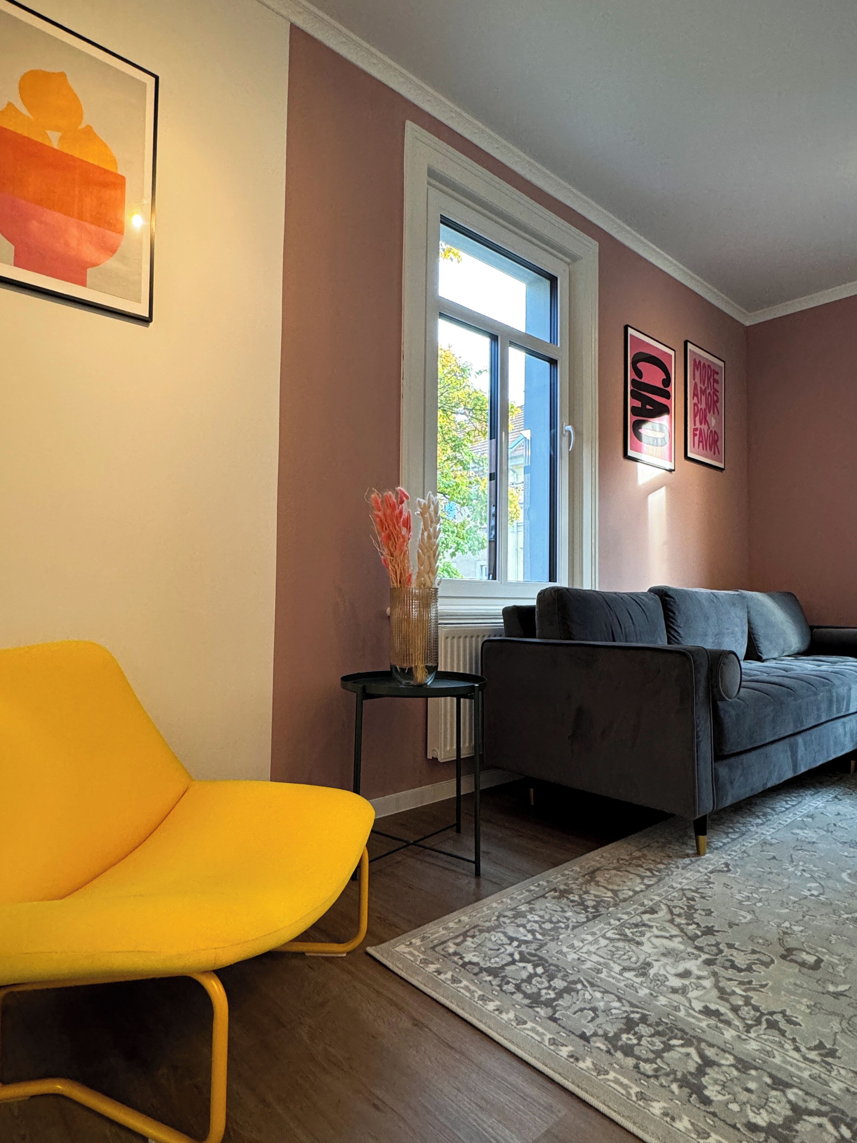 A living room with a yellow chair, a grey sofa, a patterned rug, and two framed pictures on the wall.