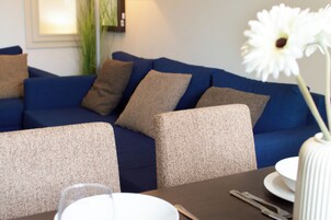 A modern living room with a blue sofa, beige cushions, and a dining table with a white vase and a bowl.