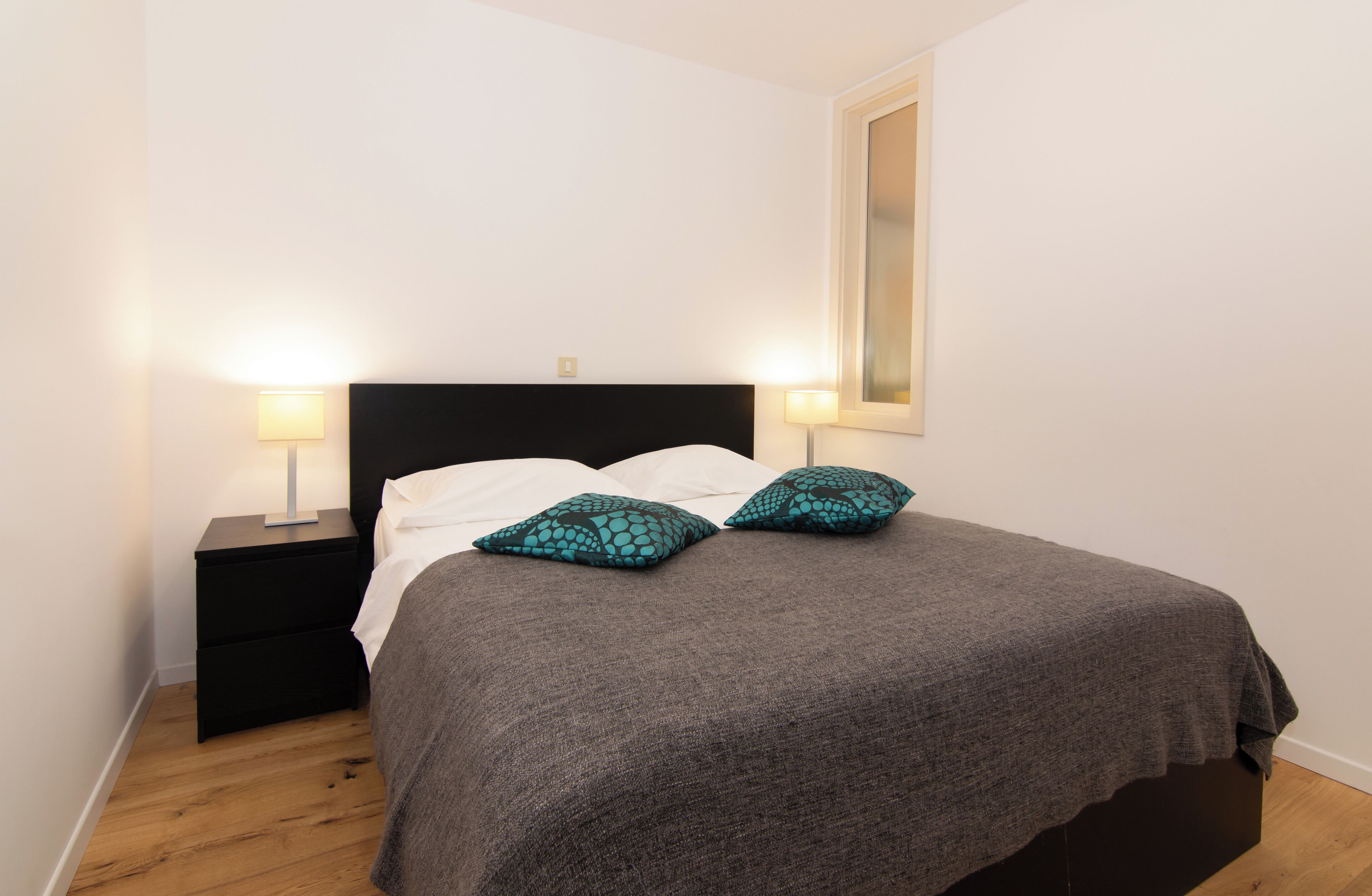 A neatly made bed with a grey blanket and two teal patterned pillows, flanked by two bedside tables with lamps, in a room with wooden flooring and white walls.